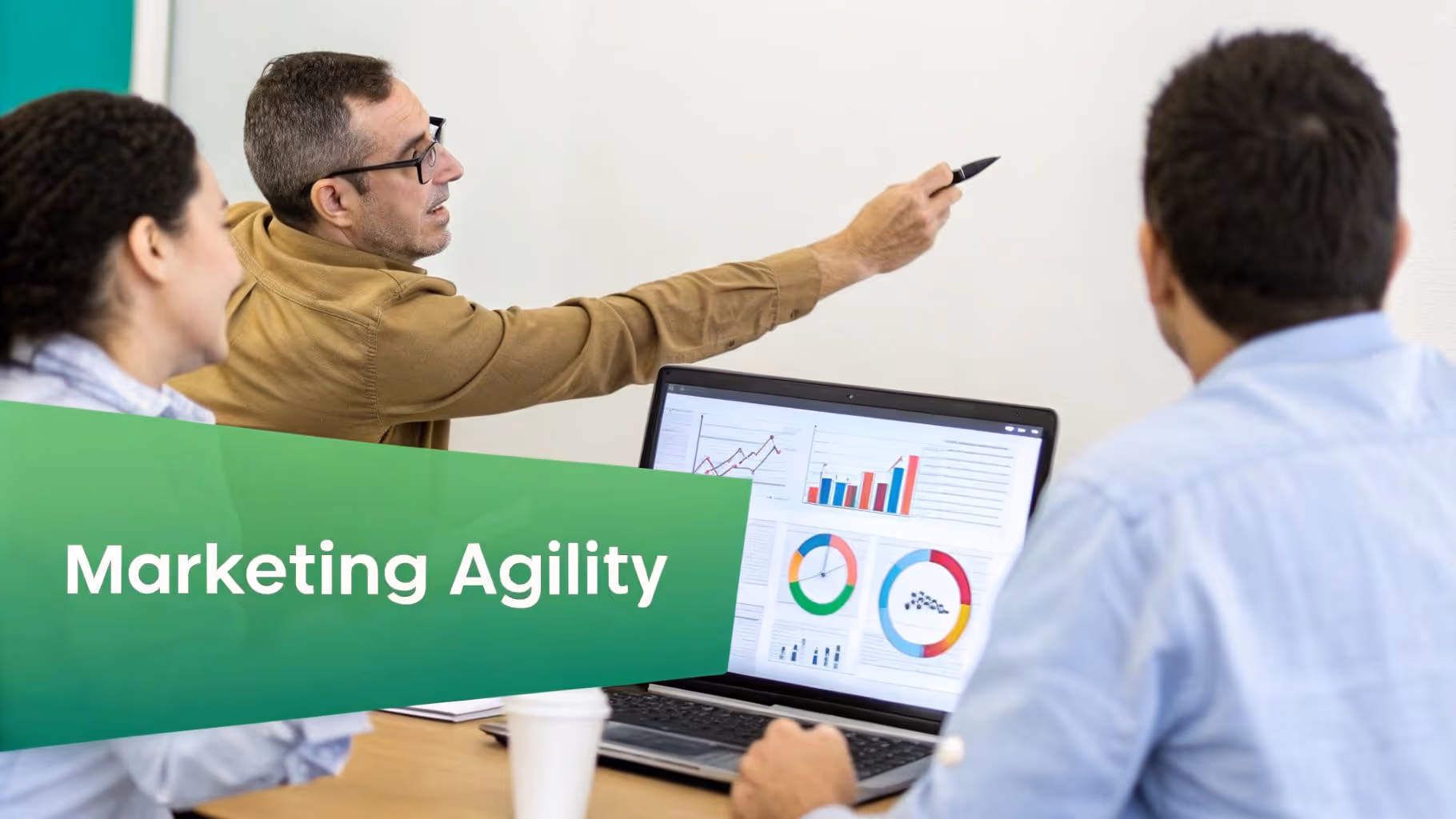 Three professionals discuss marketing agility in an office, with one presenting and another analyzing data on a laptop.